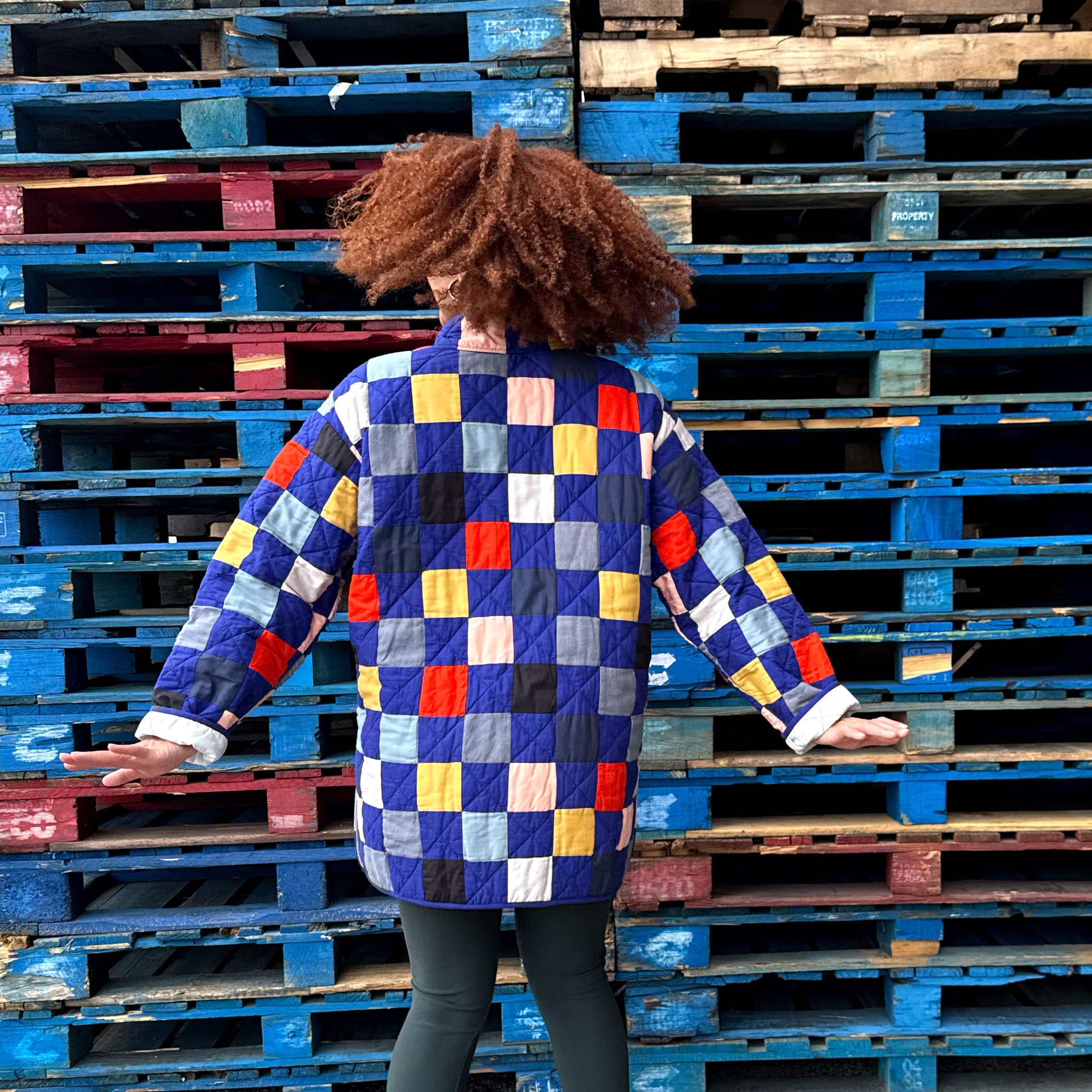 Person wearing a colorful blue checkered quilted jacket standing between two stacks of pallets.