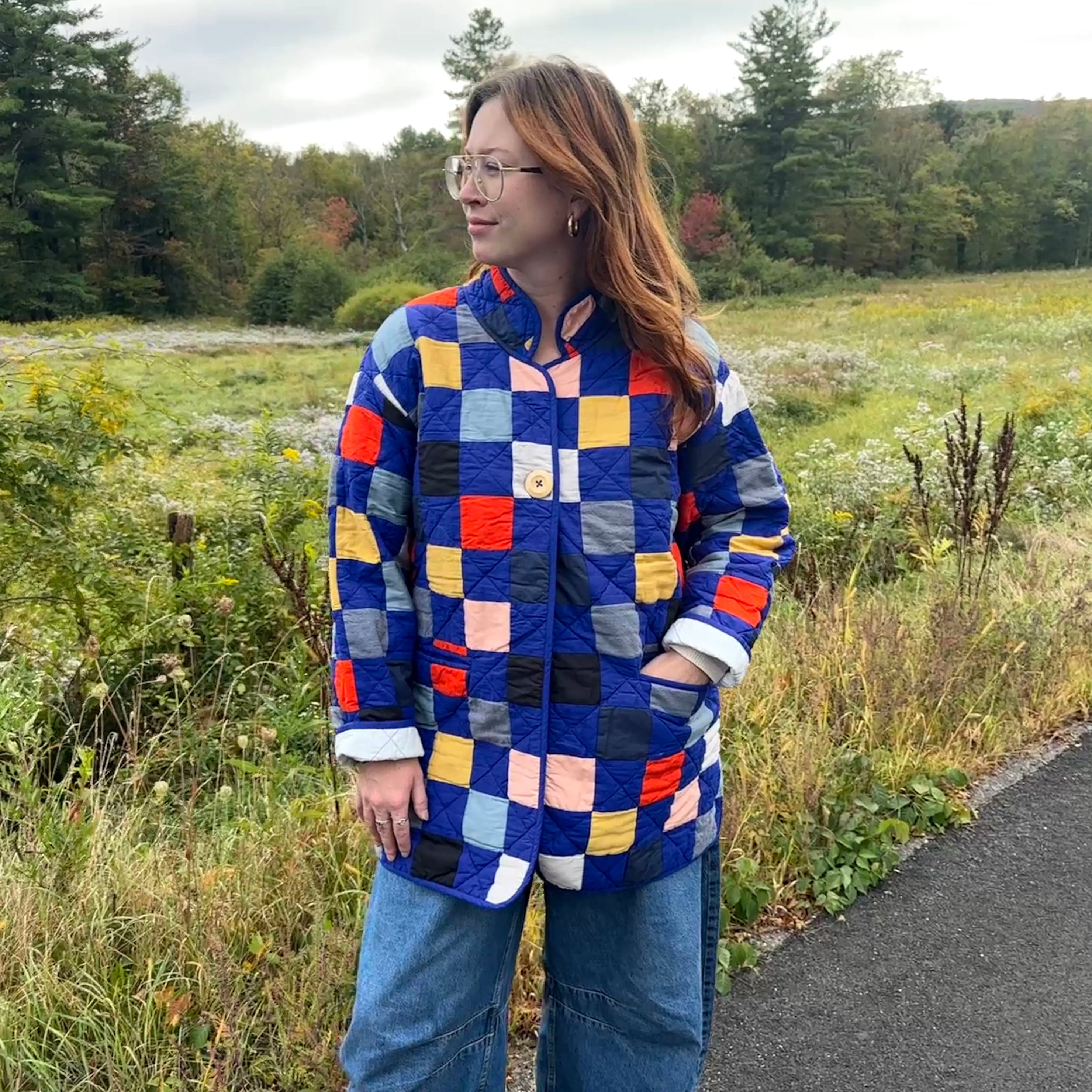 Person wearing a colorful checkered blue quilt coat in a natural setting with trees and grass.