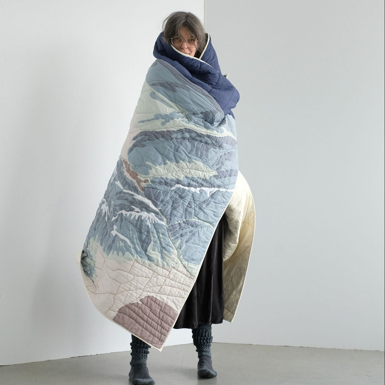 Person wrapped in a large, textured Wasatch Range quilt against a plain white wall.