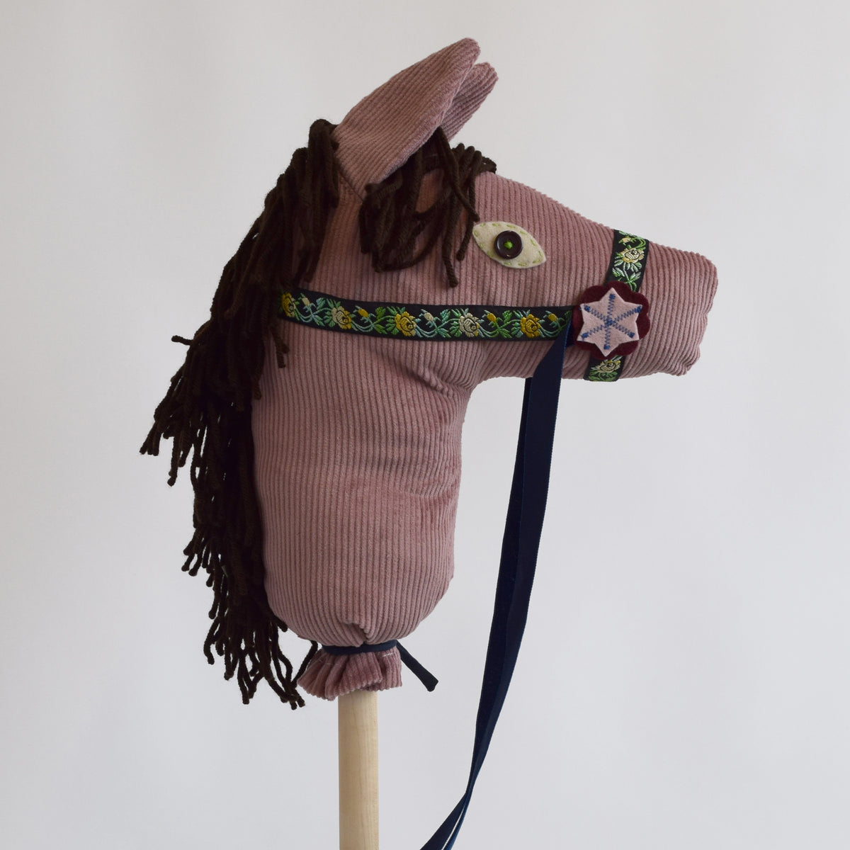 A handmade pink corduroy hobby horse, featuring a dark brown yarn mane, embroidered bridle details, and a dark blue strap, photographed against a plain light gray background.
