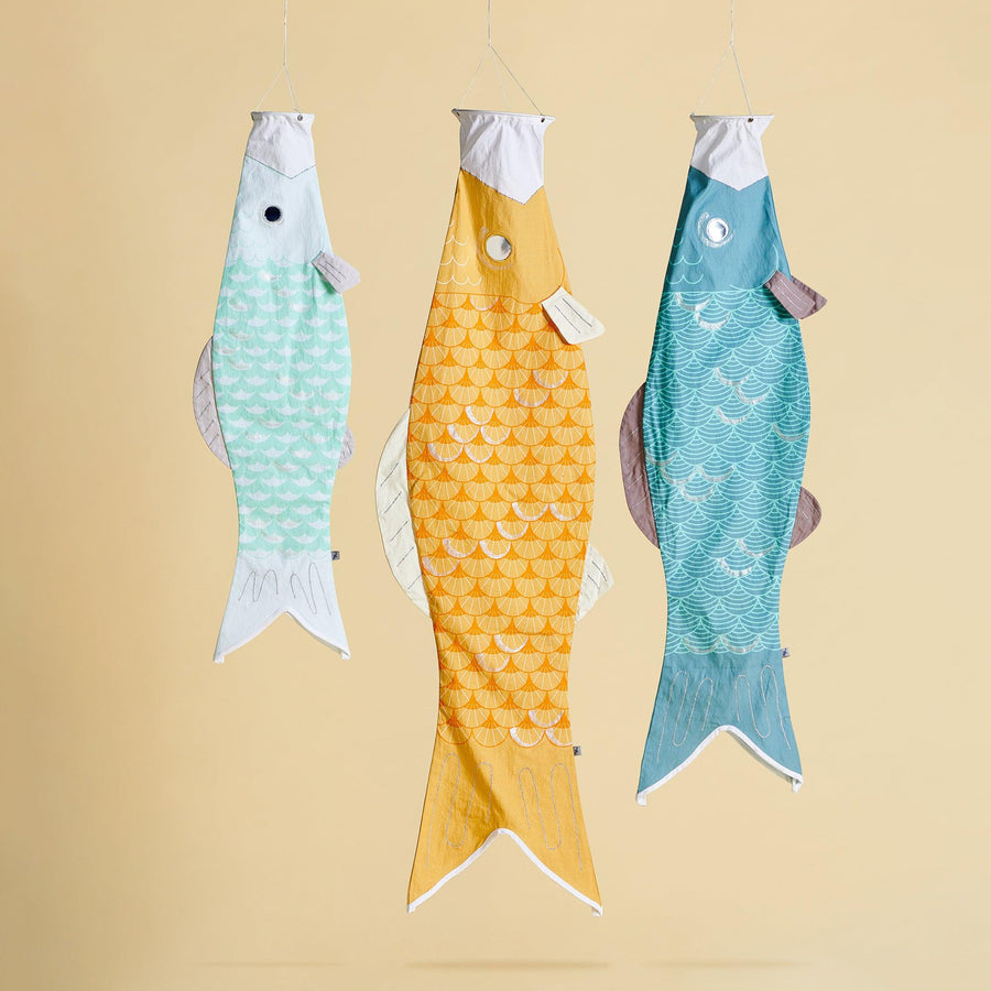 Three windsocks hanging on a yellow background. A small light blue fish is on the left, a large yellow windsock fish is in the middle and a medium size fish is on the end. 