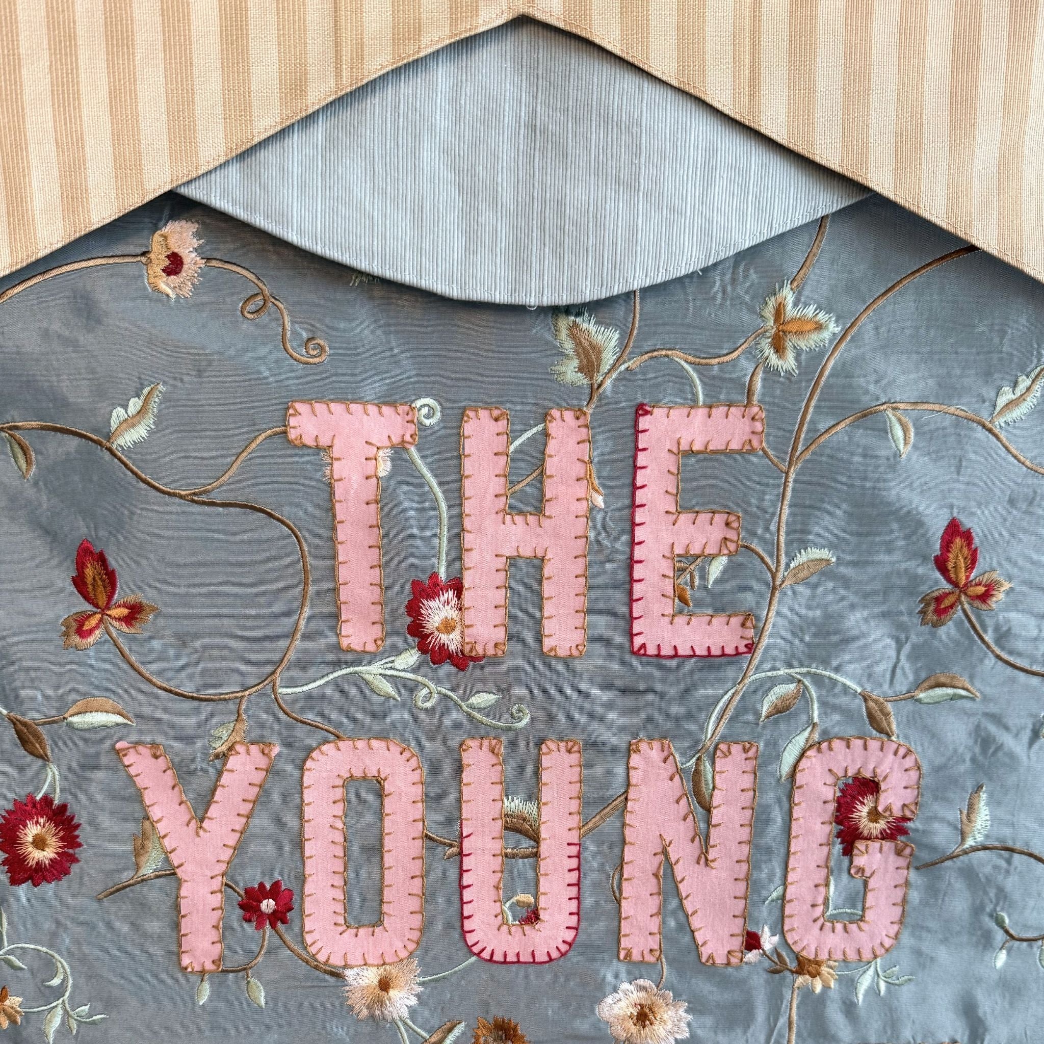 Decorative fabric with embroidered text 'THE YOUNG' surrounded by floral patterns on a textured background.