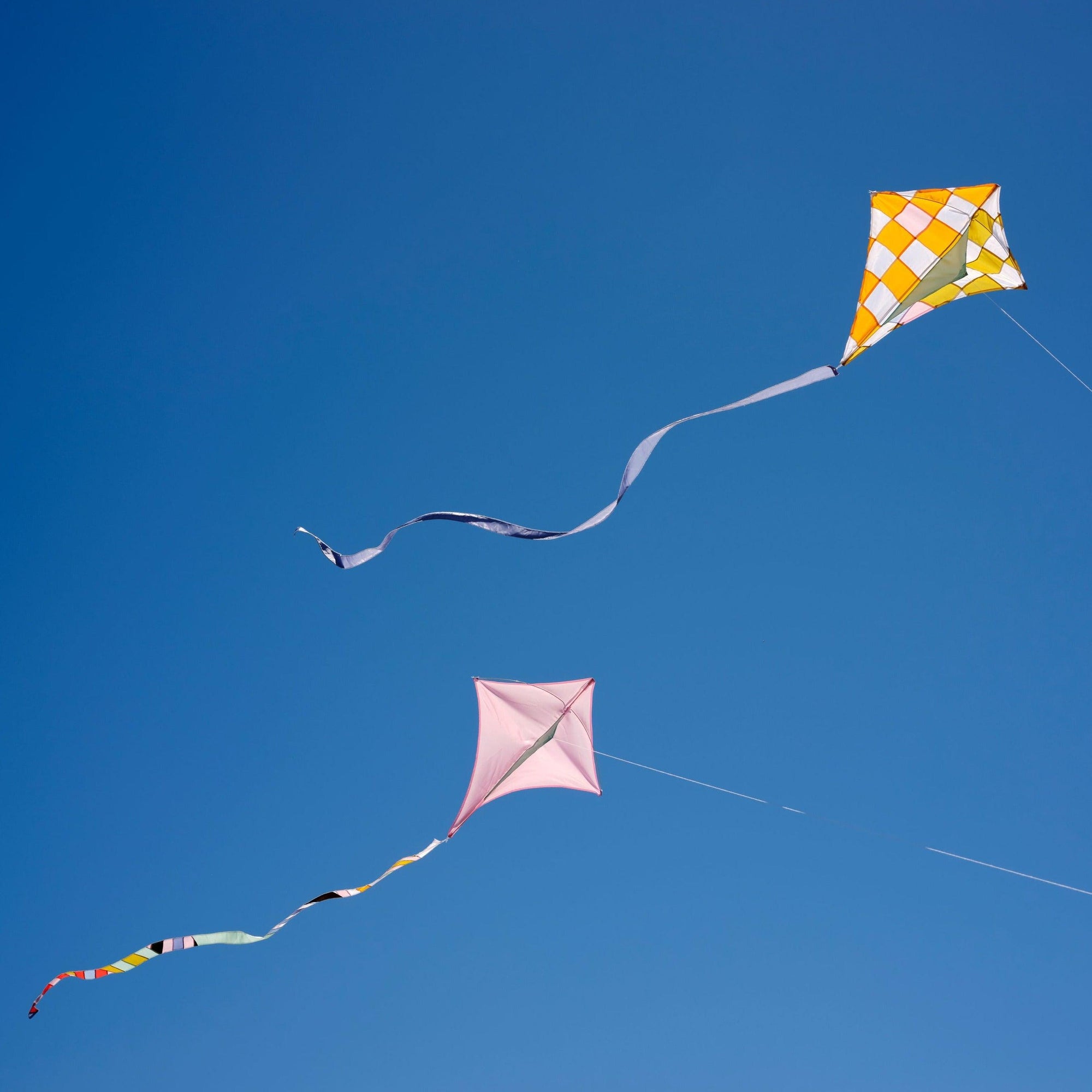 A rose colored diamond kite and a yellow checkered patchwork kite flying high in the blue sky_Haptic Lab