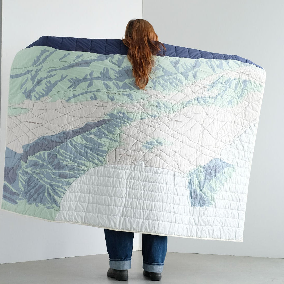 Person holding a large quilt with a Los Angeles landscape design against a white wall.