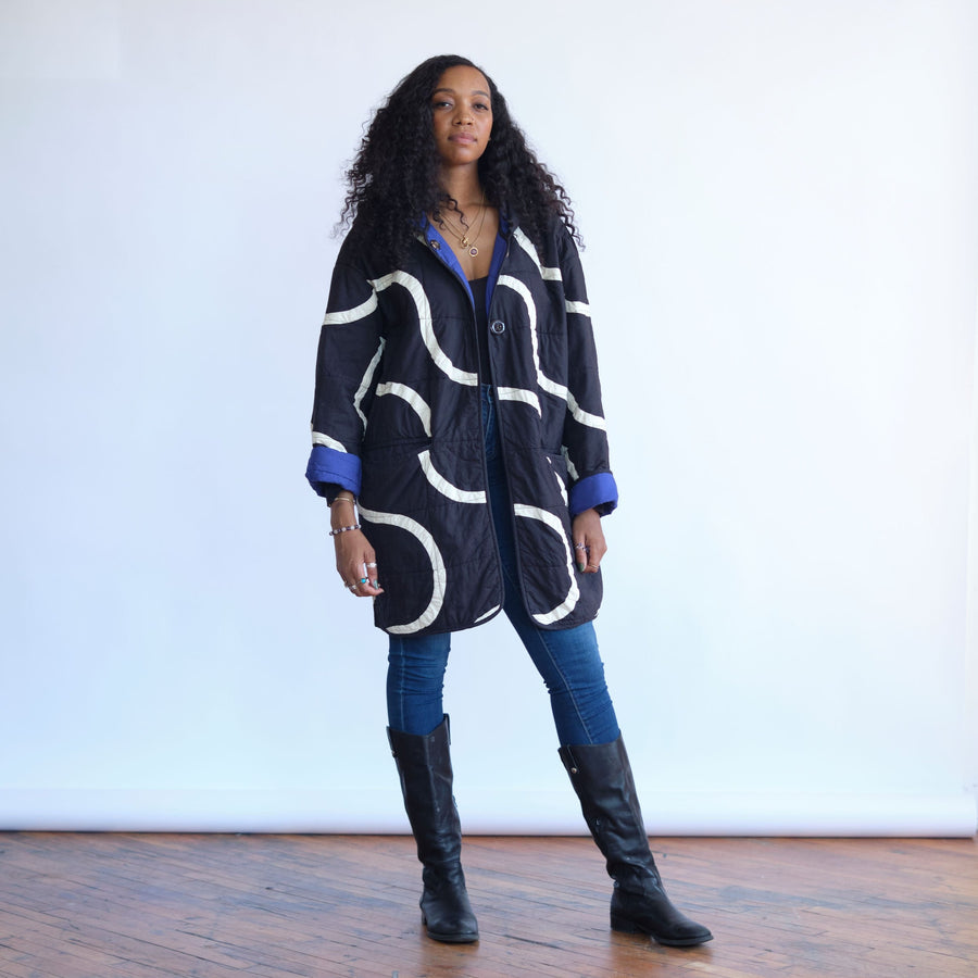 Snake Path Quilt Coat by Haptic Lab. Black quilt coat with white patchwork snake design. Sleeves rolled up to show blue interior of the coat. 