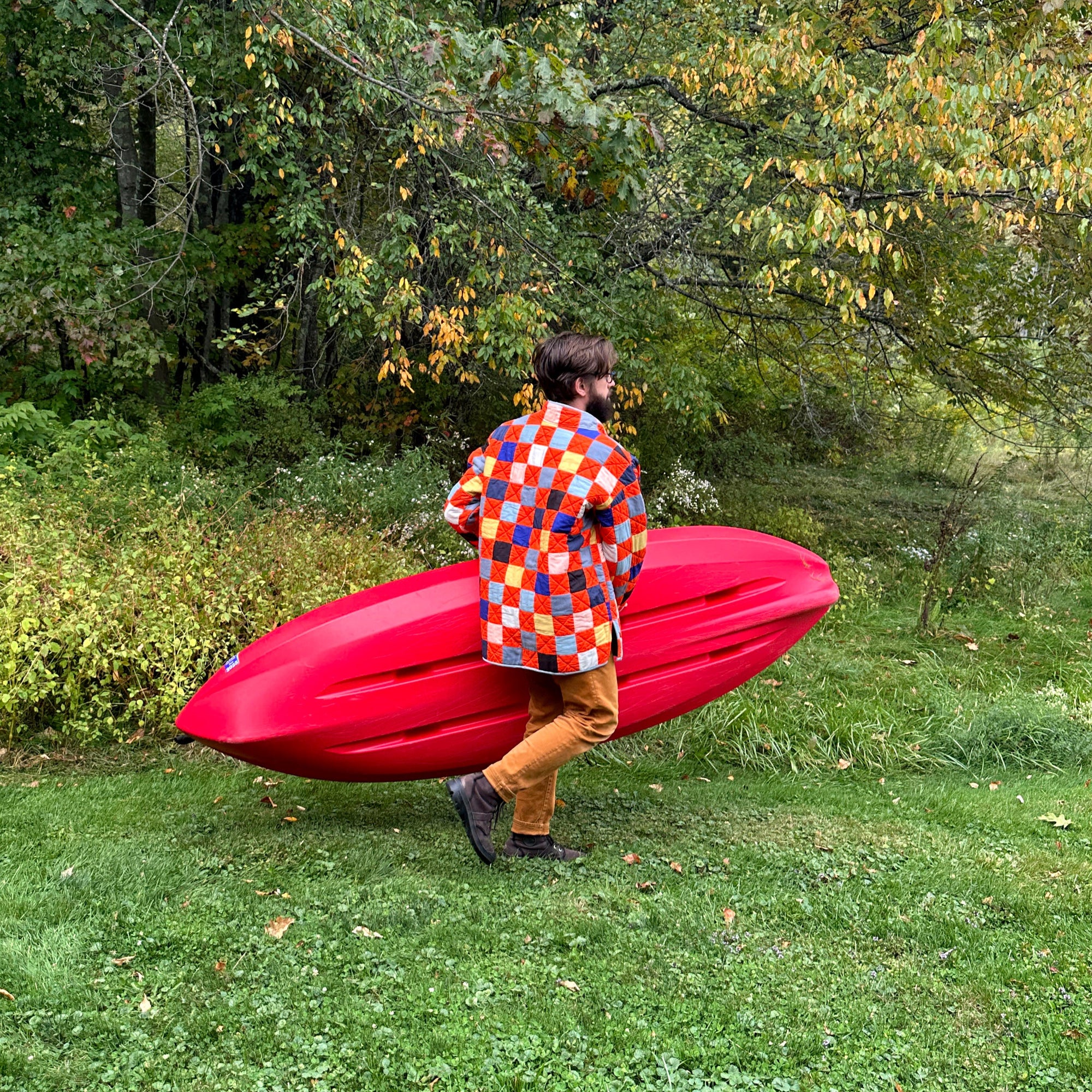 Person in a red quilt coat holding a red kayak in a forested area