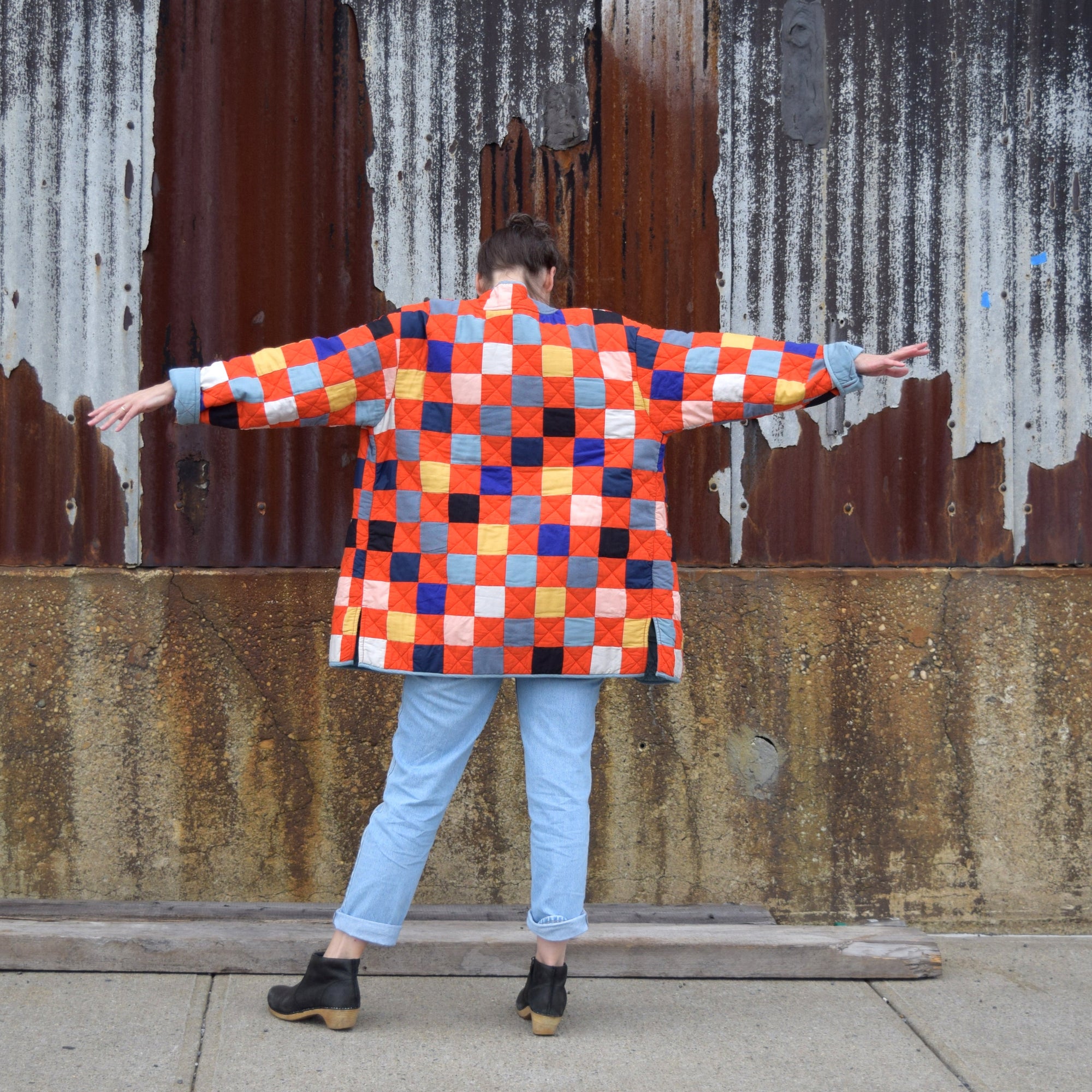 Person wearing a red patchwork quilted jacket standing in front of a rusty metal wall.