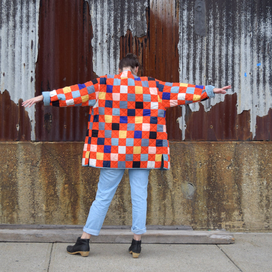Person wearing a red patchwork quilted jacket standing in front of a rusty metal wall.