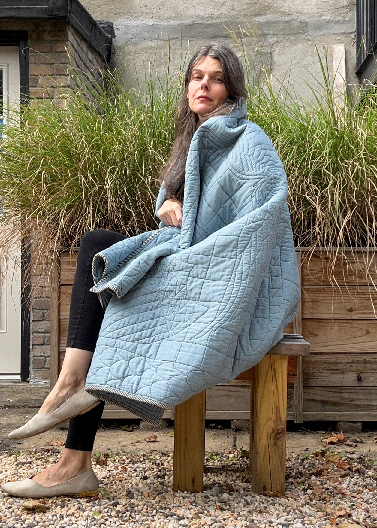 Woman sitting outdoors with a Haptic Lab quilt draped over her, surrounded by tall grass and a wooden bench.
