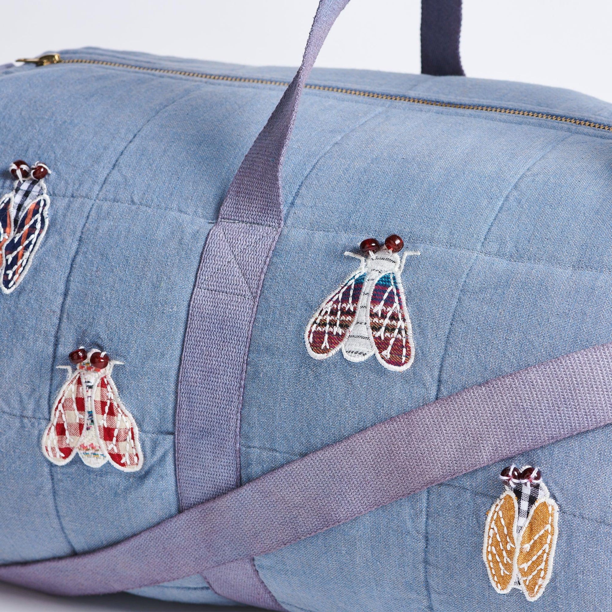 Close up picture of light Blue Duffle  bag with appliquéd cicada's on a white background. Cicadas are appliqued with remnant fabrics and wing details are embroidered. 