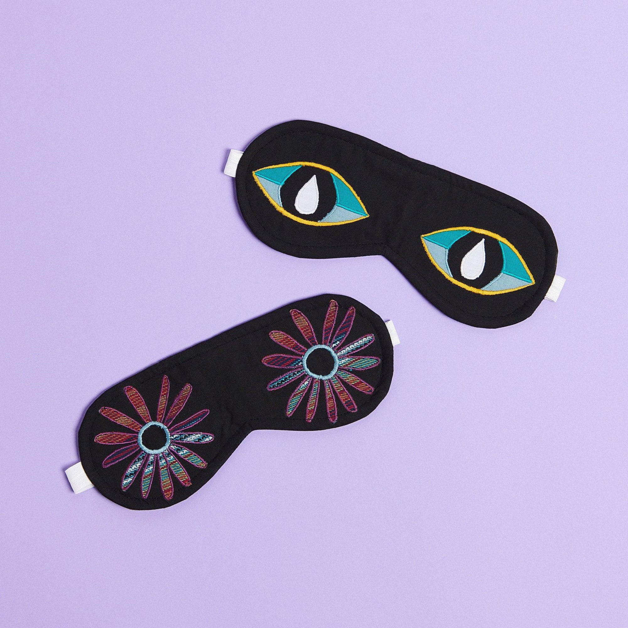 Two eye masks on a purple background. Eye Mask with Flower eyes made out of a woven purple blue green fabric and and eye mask with wide awake eyes. The eyes are teal blue on top, light blue on the bottom and a black pupil with a teardrop in the center. 