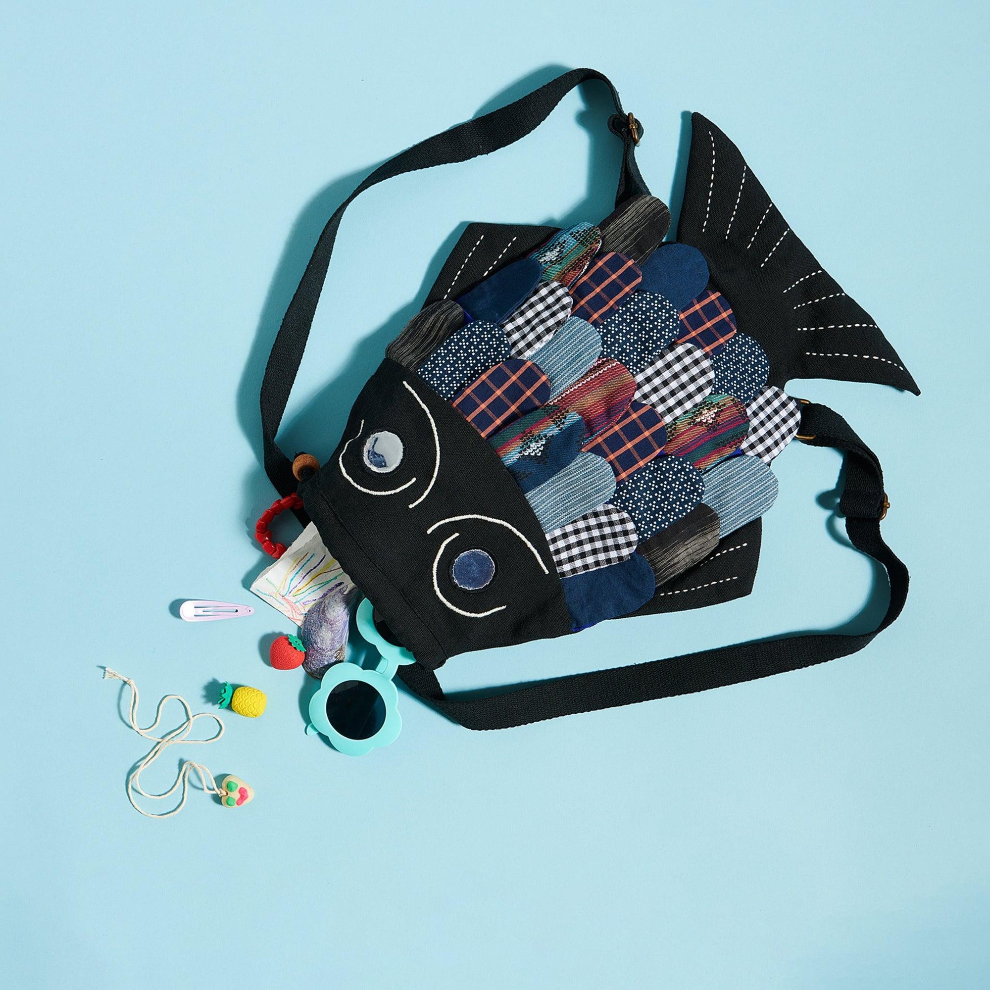 Black fish shaped backpack on a blue background. Fish has mettalic eyes and scales made out of patchwork fabrics