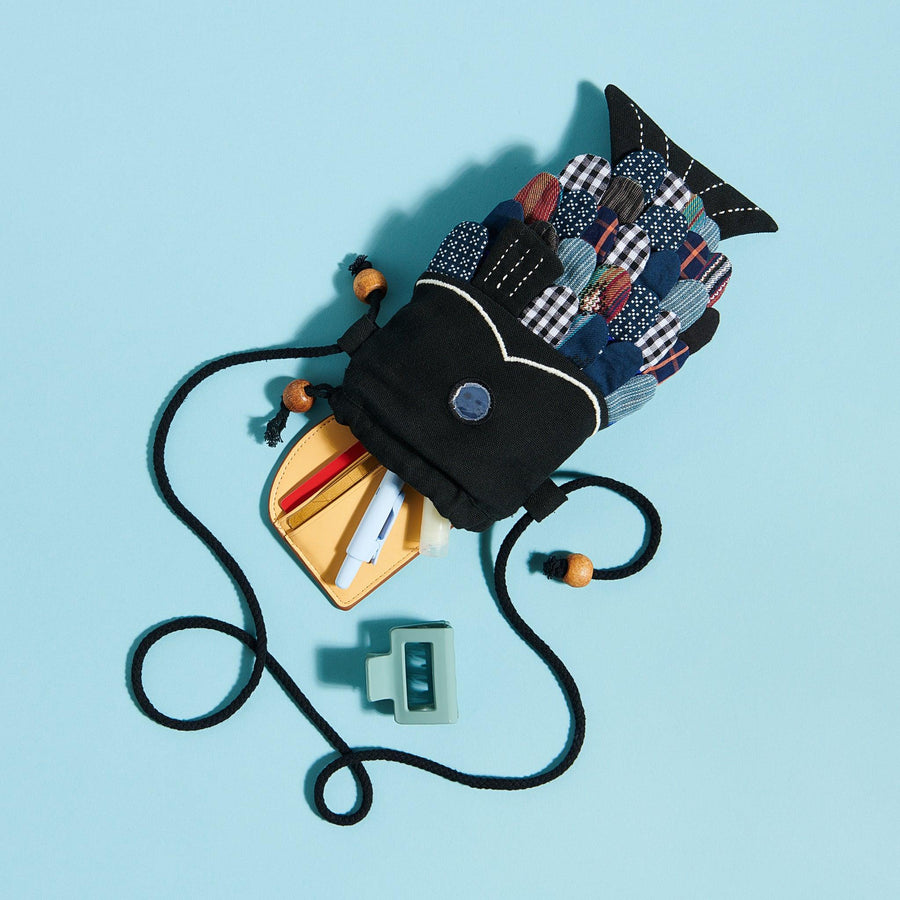 Black fish purse with patchwork scales on blue background with pens and wallet coming out of the mouth of the fish. 
