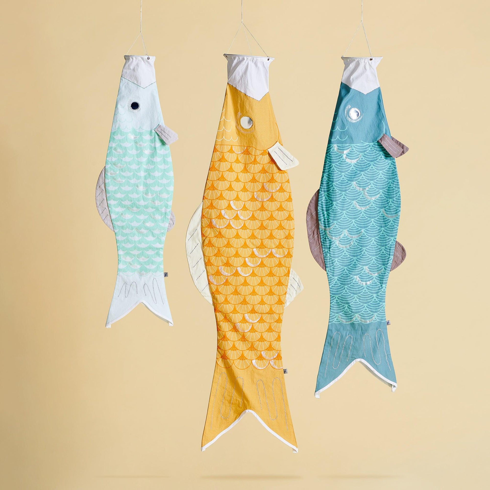 Three windsocks hanging on a yellow background. A small light blue fish is on the left, a large yellow windsock fish is in the middle and a medium size fish is on the end. 